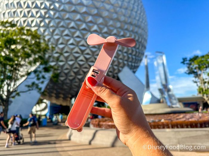 A handheld JISULIFE fan, a portable personal cooling device, is shown in the foreground with the iconic Spaceship Earth geodesic sphere at Epcot in Walt Disney World in the background.